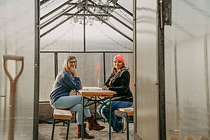Nestbau-Koordinatorin Helen Bauer und Zuzüglerin Becky Hellwig im Gespräch. Dabei sitzen sie in einem Gewächshaus, welches als Sitzecke mit Sand unter den Füßen umfunktioniert wurde.