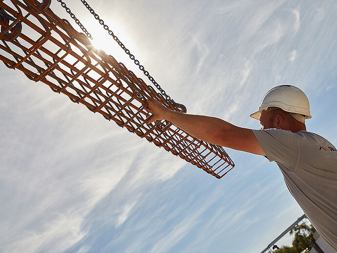 Ein Bauarbeiter überwacht den Transport von Stahl, der an einem Kran hängt.